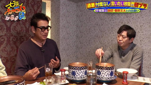 二人の男性が飲食店で鍋料理を囲んでいる様子です。一人は眼鏡をかけ話しており、もう一人は箸で料理を取っています。テレビ番組「大阪おっさんぽ」の一場面で、宇梶剛士と菅原文太の出会いについて語られているようです。