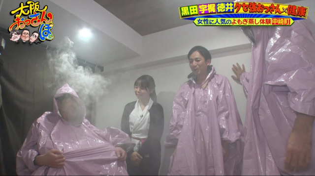 ピンクのポンチョを着た男性たちが、女性に人気のよもぎ蒸しを体験している様子。蒸気が立ち込める中で、健康と美容のためのリラックスタイムを楽しんでいるバラエティ番組の一場面です。