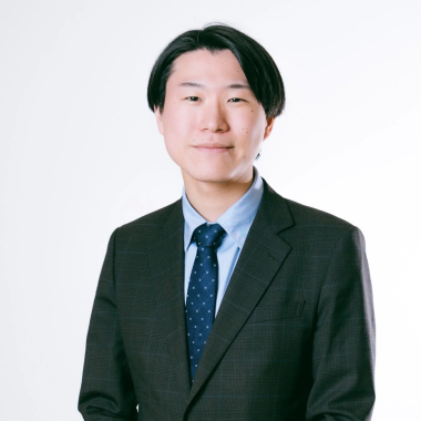 白い背景の前で、ダークカラーのスーツと水色のシャツ、ドット柄のネクタイを着用した若いアジア人男性のポートレートです。男性はカメラに向かってわずかに微笑んでおり、プロフェッショナルな印象を与えます。