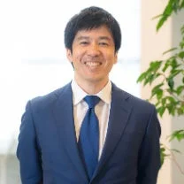 青いスーツとネクタイを着用したアジア系の男性が、カメラに向かって笑顔で立っているポートレート写真です。ビジネスシーンやプロフィール写真に適した清潔感のある印象です。