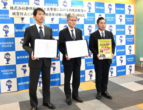 静岡県警察と株式会社静岡新聞が特殊詐欺被害防止に向けた情報啓発活動に関する協定締結式を行っている写真です。スーツを着た3人の男性が書類や啓発ボードを手にしています。