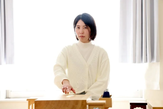 白いカーディガンを着た女性が、明るい光が差し込む室内で木製のテーブルの前に座っています。穏やかな表情で、何らかの伝統的な活動を行っているように見えます。